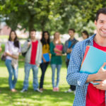 College boy holding books with blurred students in park